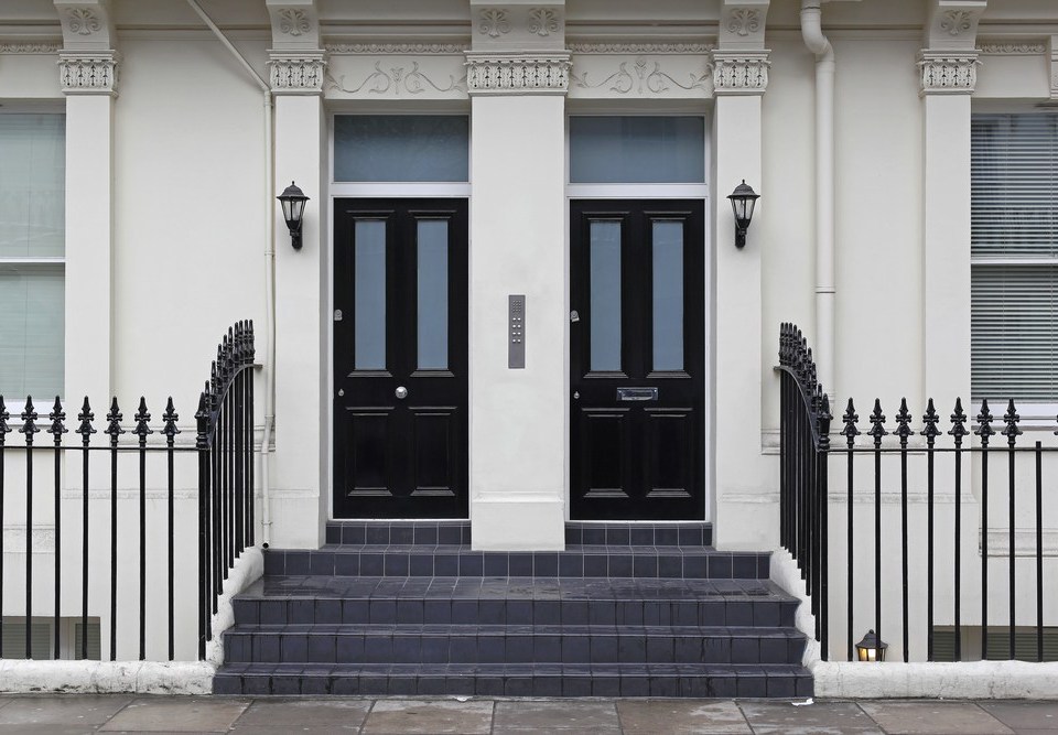 A photograph of the front of an off white building with two black front doors and steps leading up to them
