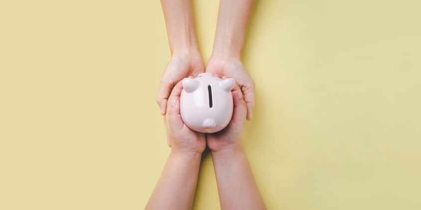 Childs hands holding a piggy bank with an adults hands underneath the childs hands.