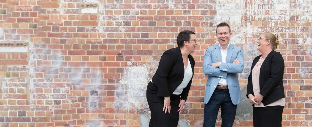 Dan, Kate and Orlena laughing together in front of a distressed brick wall.